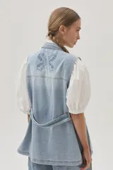 Chaleco largo de jean celeste con cuello camisero, sin mangas, con bolsillos frontales, laterales e internos, y lazo ajustable en la cintura.