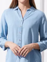 Camisa gris de tencel con cuello con solapa, escote en V y abertura central con botones. Largo de manga regulable con presilla y botón. Ruedo en espalda con tajo.