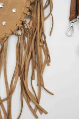 Cartera de gamuza color camel con diseño semicircular, decorada con flecos largos en el borde y tachas metálicas plateadas en la parte frontal. Incluye una correa larga ajustable y extraíble.