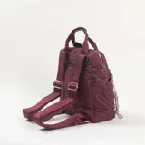 Mochila de diseño acolchado en color bordó, con costuras decorativas que forman letras. Cuenta con un compartimento principal con cierre, un bolsillo frontal con cierre, asas superiores de mano y correas ajustables. Incluye un colgante decorativo con cuentas en el cierre frontal.