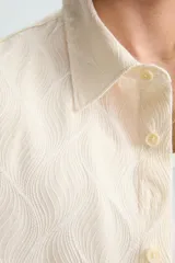 Camisa de corte relajado (relaxed fit) color crema o blanco roto, confeccionada en tejido con mezcla de algodón. Presenta un bordado sutil en relieve con patrón de ondas o líneas diagonales entrecruzadas. Tiene cuello solapa, manga corta y un bolsillo de plastrón en el pecho.