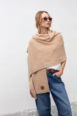 Chal de tejido de punto en color beige, con textura suave y diseño envolvente. Presenta una etiqueta de cuero sintético con logo grabado en la parte inferior.
