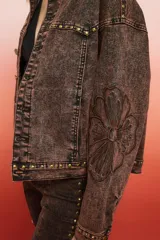 Campera de jean con lavado oscuro rojizo/marrón, estilo trucker. Presenta tachas doradas a lo largo de los bordes de los bolsillos, el canesú y los puños. Posee un bordado en relieve de diseño floral en la manga.