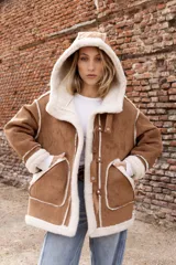 Campera estilo gamulán color tostado con interior y bordes de corderito sintético color crema. Presenta capucha amplia, cierre frontal con trabas y broches metálicos, y bolsillos delanteros grandes. Posee un calce relajado y diseño de textura suave.