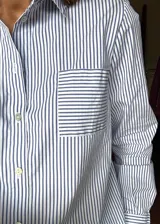 Camisa de popelina a rayas verticales blancas y azules, de estilo clásico con cuello, bolsillo frontal con rayas en dirección horizontal, cierre delantero con botones de nácar y mangas largas con puños.
