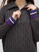 Pullover de algodón color beige, con tejido en ochos, cuello polo con cierre frontal y puños a rayas de colores (blanco, bordó y amarillo). El diseño es holgado y cae por debajo de la cadera.