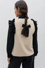 Chaleco de lana color beige con bordados florales artesanales en tonos marrones y blancos. Presenta un diseño sin mangas con volados en los hombros y cierre frontal mediante lazos negros ajustables.