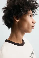 Camiseta de corte relajado confeccionada en algodón, color blanco crudo con cuello redondo en contraste color negro. Presenta un bordado frontal con la frase 'Coração de Melão' y un parche en forma de corazón.