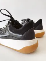Zapatillas deportivas negras de cuero, con cordones y suela de goma blanca con base marrón.