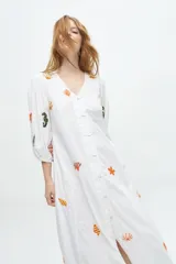 Vestido largo blanco de algodón con bordados de motivos marinos en tonos naranja, verde y azul. Tiene escote en V, mangas 3/4 abullonadas y cierre frontal con botones.