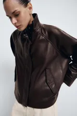 Chaqueta bomber de piel color marrón oscuro, con cuello alto, cierre frontal con cremallera y solapa con botones ocultos. Presenta mangas largas, bolsillos delanteros y un diseño de corte recto.