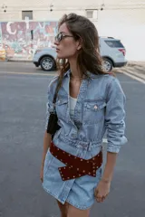 Vestido corto de denim azul claro, estilo camisa, con cuello solapa, escote pico, manga larga y cierre frontal de botones metálicos. Presenta bolsillos delanteros con tapa y bolsillos de plastrón en la espalda. La cintura está ajustada con elástico fruncido.