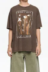 Camiseta de algodón orgánico color marrón oscuro con efecto gastado, corte maxi oversize y manga corta ancha. Presenta un estampado central enmarcado con motivos florales y el texto "BIMBA Y LOLA" en blanco.