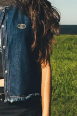 Chaleco de denim azul oscuro con costuras contrastantes en hilo ocre, cierre frontal con broches metálicos a presión y detalle de bordado ovalado en el pecho.