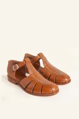 Sandalias de cuero color marrón, tipo romanas, con talón cerrado, tira en T con detalles de tachas metálicas, aberturas laterales y cierre de hebilla ajustable. Tienen tacón bajo de madera.