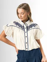 Blusa blanca de algodón con cuello mao, mangas globo cortas y cierre frontal con botones. Presenta un canesú destacado con bordados en azul marino con detalles calados.