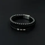 Pulsera negra de cuatro tiras: una de cuero trenzado, una de cuero liso con tres cilindros metálicos y una de cuentas esféricas metálicas.