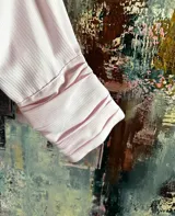 Camisa de algodón color rosa suave con textura de rayas verticales sutiles. Presenta un cuello grande con volantes que se extiende hacia el frente y mangas largas con puños abotonados.