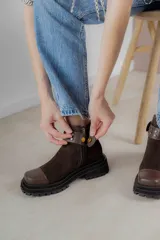 Bota corta de gamuza marrón con puntera de cuero liso, suela track negra y correa desmontable con tachas metálicas.