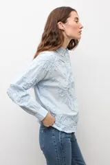Camisa celeste con apliques florales bordados, cuello camisero, manga larga y cierre frontal de botones.