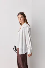 Camisa blanca de manga larga con textura suave y caída liviana, cuello camisero, puños anchos y fruncidos sutiles en los hombros.