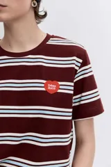 Remera de manga corta con cuello redondo, confeccionada en tejido de punto con diseño de rayas horizontales en tonos marrón, blanco y celeste. Presenta un pequeño parche circular rojo en el pecho.