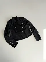 Campera de estilo militar en color marrón oscuro, confeccionada en gamuza. Presenta un diseño cruzado con doble fila de botones metálicos grabados, cuello alto y charreteras en los hombros.
