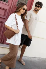 Cartera tipo bandolera de color negro, confeccionada en cuero sintético con acabado aterciopelado. Cuenta con una correa larga ajustable decorada con remaches metálicos circulares.