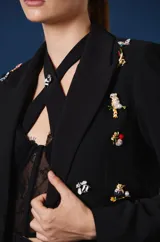 Saco tipo spencer negro de corte corto y estructurado, con solapas clásicas y detalles de pines decorativos distribuidos en la parte frontal.