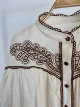 Blusa off-white de algodón con canesú de broderie en tono chocolate, cuello mao, mangas globo y cierre frontal con botones.