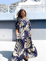 Vestido largo estilo túnica con cuello camisero y botones frontales, mangas tres cuartos y falda con volados en capas. Presenta un estampado grande de hojas tropicales en azul marino y crudo.