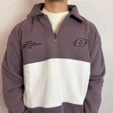 Buzo de algodón con medio cierre frontal y cuello tipo polo. Presenta un diseño de bloques de color en tonos gris oscuro y blanco, con bordados decorativos en el pecho.