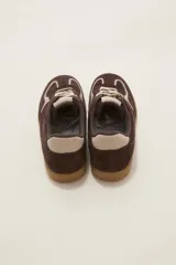 Zapatilla urbana de gamuza color marrón con detalles en beige, cordones claros y suela de goma tipo caramelo.