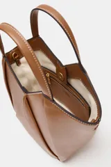Bolso mini shopper de cuero sintético en color marrón, con diseño estructurado y costuras decorativas contrastantes. Cuenta con doble asa de mano y cierre magnético.