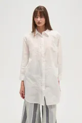 Camisa blanca de manga larga, corte holgado y largo, con cuello camisero y botones frontales. Presenta bordados florales en tono blanco sobre el tejido.
