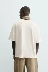 Camiseta de corte relajado (relaxed fit) color blanco roto, con cuello redondo y manga corta. Presenta un bordado sutil en el pecho con diseño floral en tonos claros.