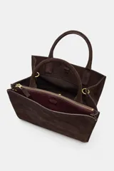 Bolso de mano formato city confeccionado en gamuza color marrón oscuro. Cuenta con doble asa de mano, asa bandolera extraíble, compartimento interior con cierre y bolsillo exterior con imán.