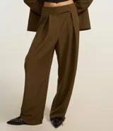 Pantalón de sastrería corte barrel, color marrón oscuro, con pliegues delanteros, cintura con cierre desplazado y piernas anchas.