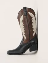 Bota texana de cuero bicolor, combinando el color negro en la base y el marrón en la caña. Presenta un diseño de apliques decorativos en color beige con forma de alas a los costados y taco cuadrado de 5 cm.