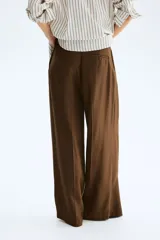 Pantalón sastrero de corte ancho, color marrón, con pinzas frontales, trabillas para cinturón y raya marcada en las piernas.