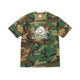 Remera de manga corta con estampado frontal que presenta una casa hecha de billetes flanqueada por dos rifles, sobre un diseño de camuflaje militar. Cuenta con el logo de la marca en la parte inferior del gráfico.