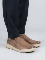 Zapato tipo slip-on Hush Puppies modelo Hunt, confeccionado en cuero color marrón con suela de EVA y goma en tono beige. Presenta elásticos laterales para facilitar el calce y un diseño de estilo casual.
