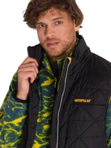 Chaleco acolchado negro con diseño de rombos, cierre frontal completo y logo de la marca Caterpillar en el pecho. Cuenta con bolsillos laterales y es resistente al agua y al viento.