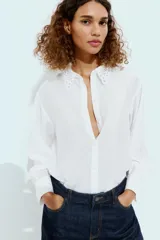 Camisa blanca de manga larga con cuello de solapa y detalles de encaje a tono en el cuello y los puños. Presenta cierre frontal con botones y corte recto.