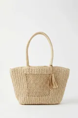 Cartera tipo shopper de mimbre tejido color beige, con doble asa de hombro y detalle de borla colgante.