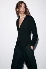 Chaqueta de punto color negro con escote en pico, manga tres cuartos y cierre frontal con botones dorados. Presenta un diseño con bajo asimétrico.