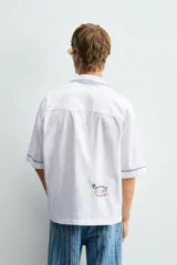 Camisa blanca de algodón con cuello bowling y manga corta, corte relajado. Presenta bordados a contraste en azul, rojo y amarillo que simulan elementos de cocina y artesanía, además de ribetes de pespunte azul en cuello, puños y abertura frontal.
