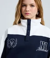 Buzo tipo polo de textura waffle con diseño color block en blanco y azul marino. Presenta cuello alto con cierre de botones, mangas largas y bordados decorativos en el frente.