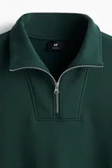 Buzo de tejido pesado color verde oscuro, con cuello alto, medio cierre metálico y hombros caídos. Presenta puños y bajo en punto acanalado.
