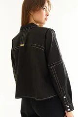 Camisa de corte oversize y estilo casual, confeccionada en algodón negro con costuras a contratono en color blanco. Presenta cuello camisero, cierre frontal parcial, mangas largas y un bordado distintivo en el puño.